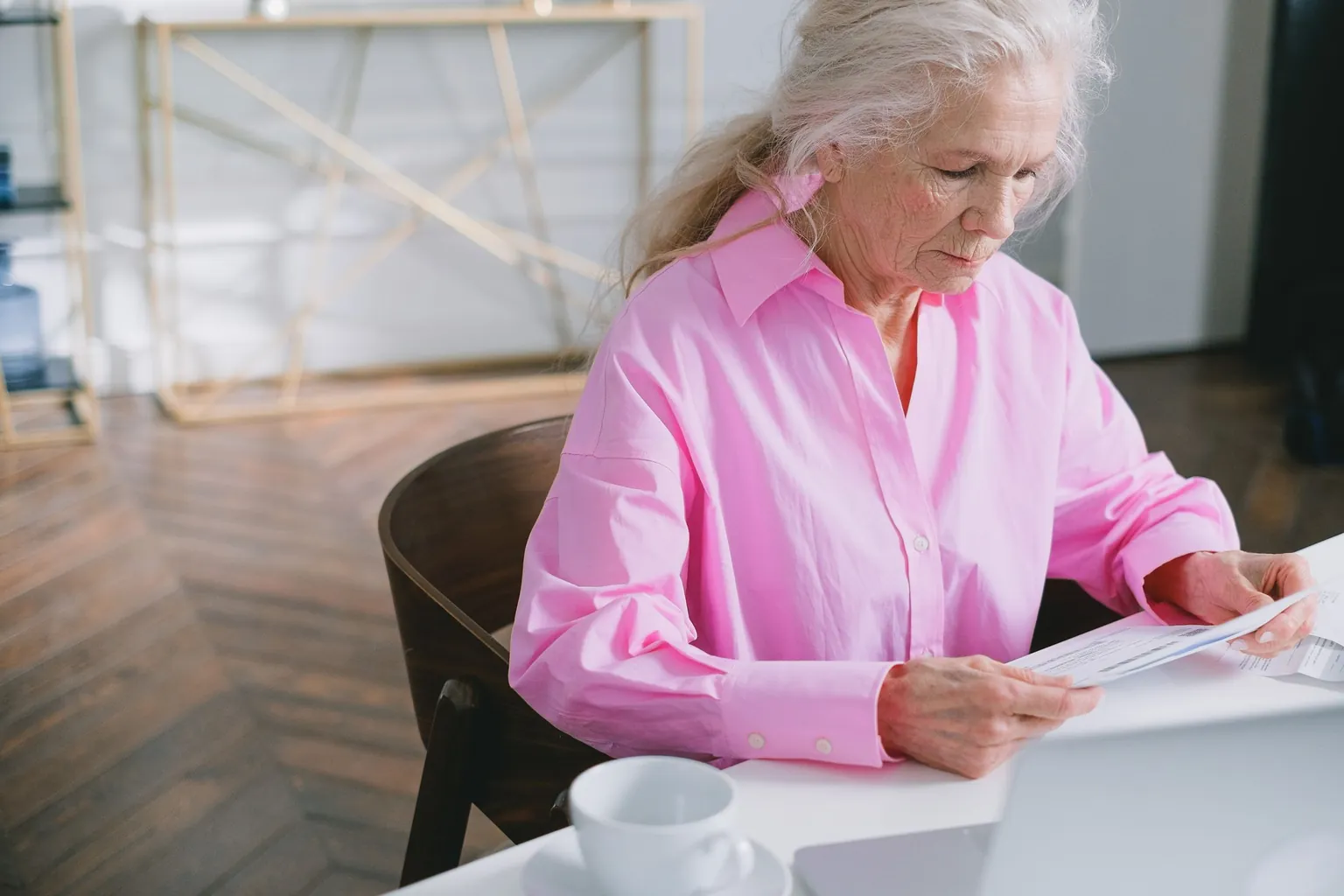 Femme aux cheveux blancs en chemise rose lisant un document papier à une table, devant un ordinateur portable et une tasse