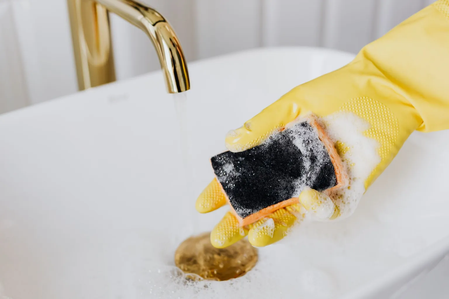 Main gantée de jaune nettoyant une vasque blanche avec une éponge savonneuse