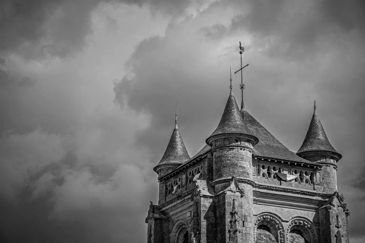 Clocher de pierre à tourelles surmonté d'une croix et d'un coq, en noir et blanc sous un ciel nuageux