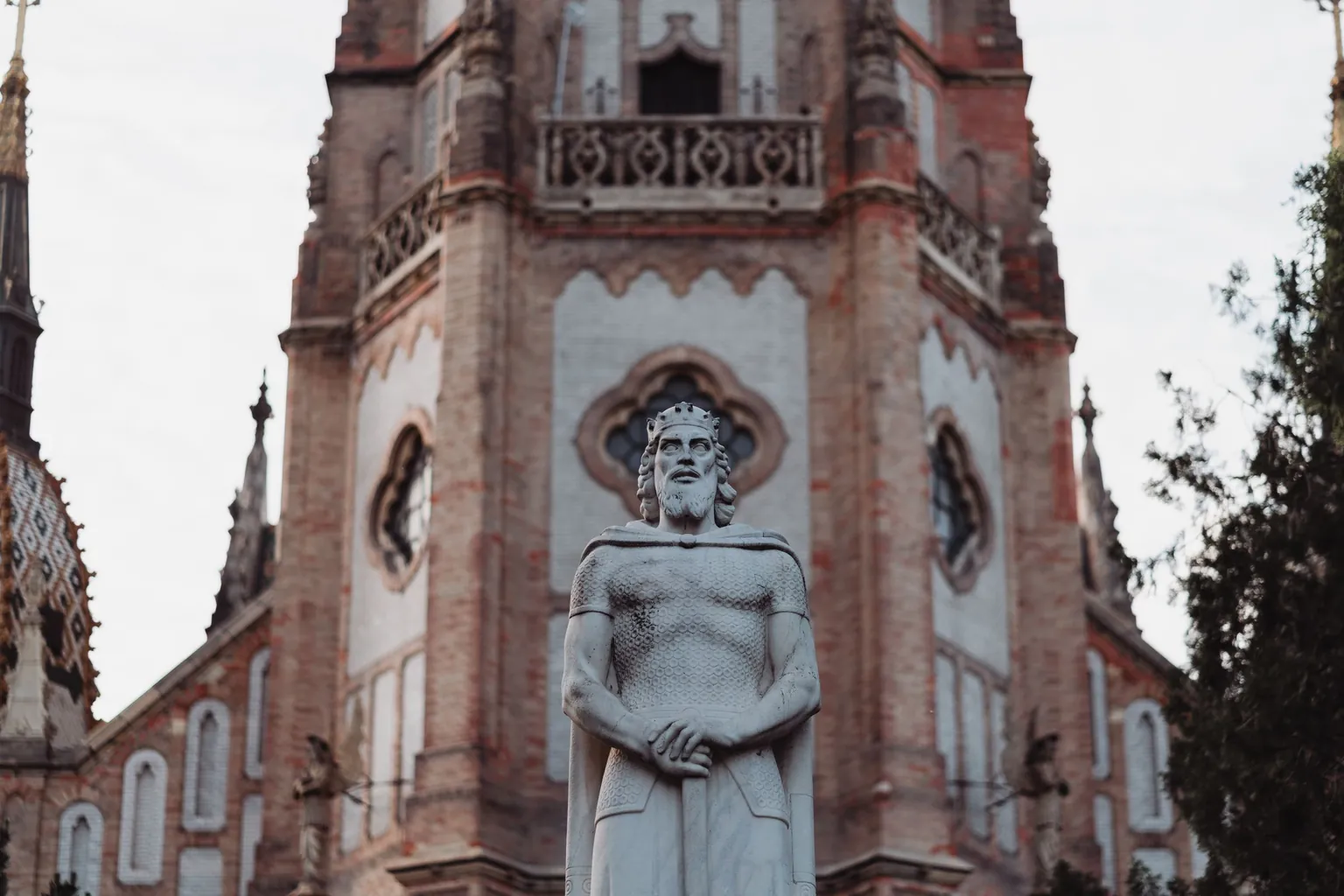 Statue de pierre d'un roi couronné en cotte de mailles, les mains posées sur une épée, devant une église néogothique en brique