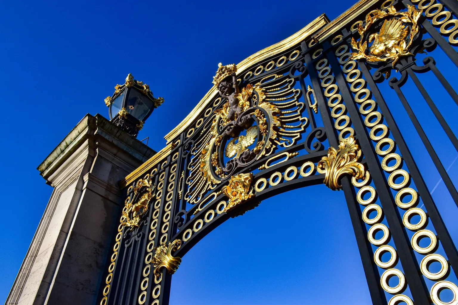 Grille monumentale en fer forgé noir rehaussée de fleurons et de volutes dorés, sous un ciel bleu