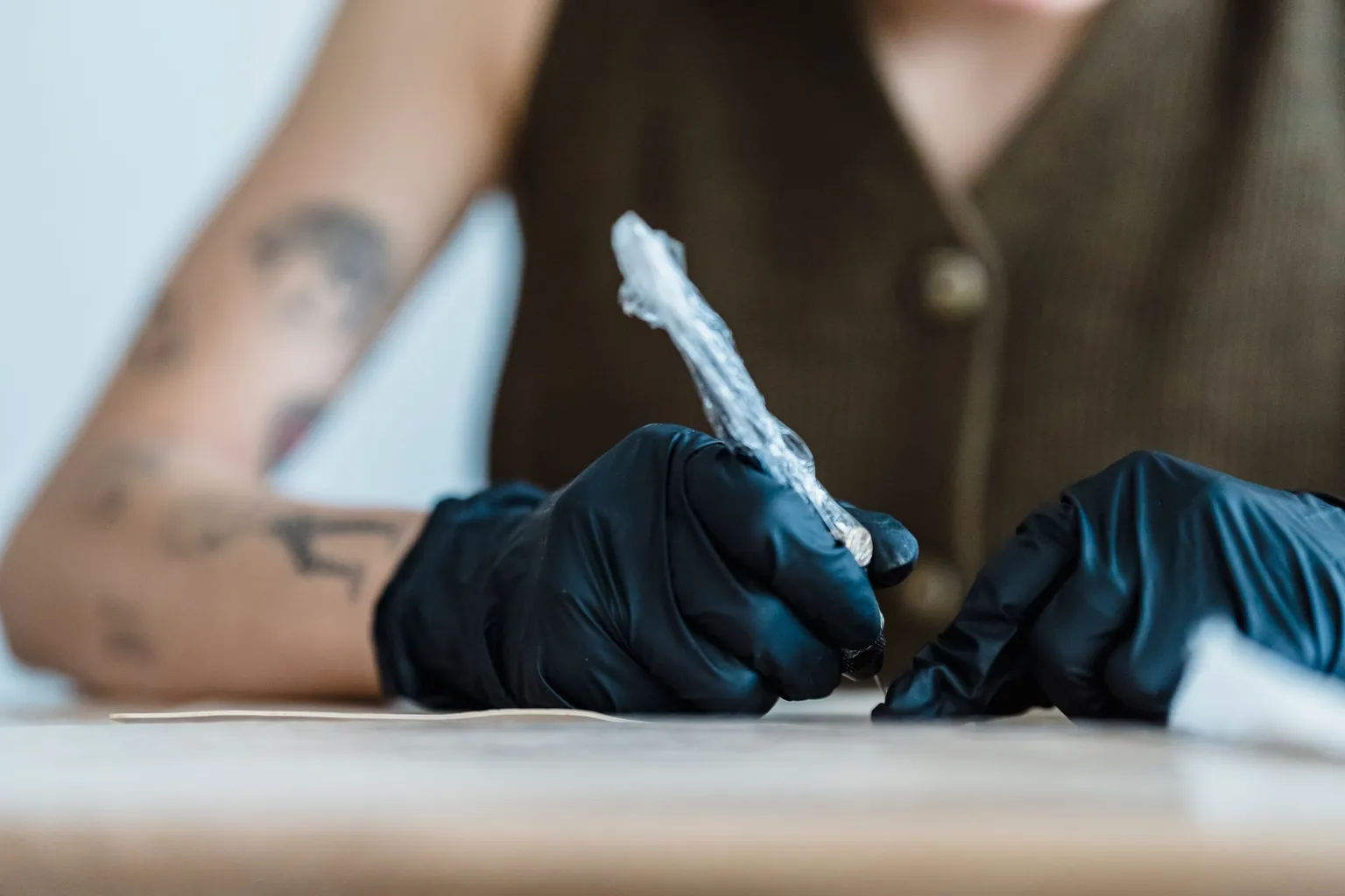 Mains gantées de noir tenant une machine à tatouer protégée par un film plastique à usage unique