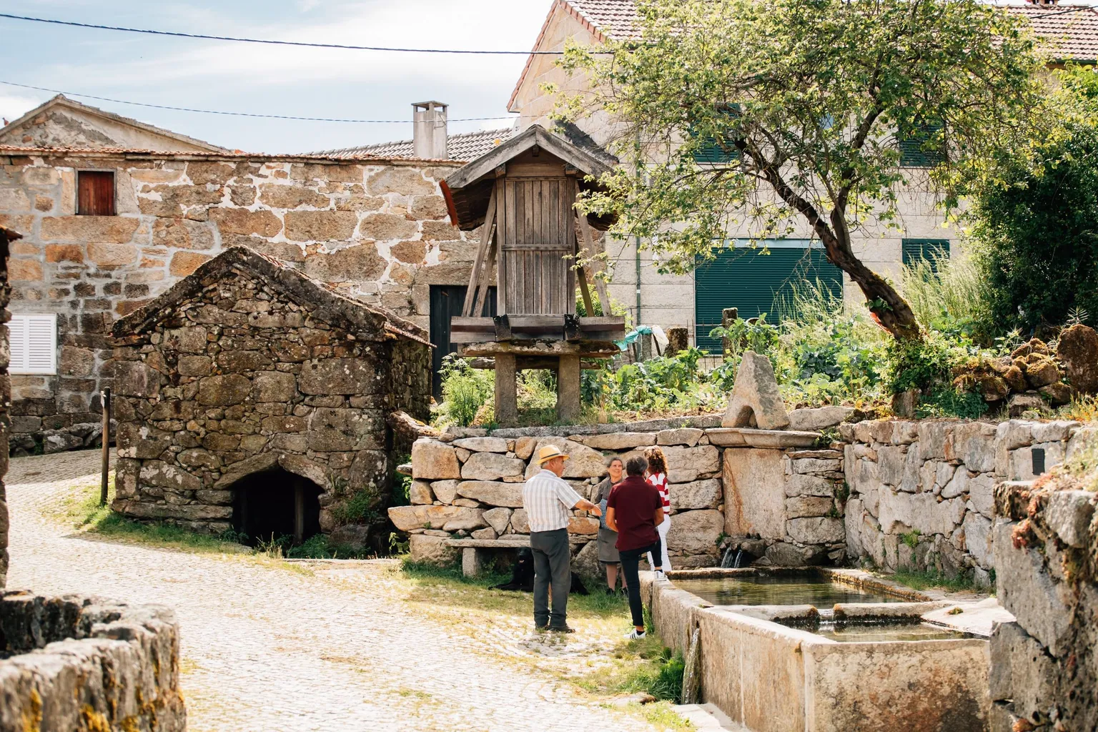 Place de village aux maisons de granit avec un grenier à grain en bois sur pilotis, une fontaine-lavoir et quelques habitants