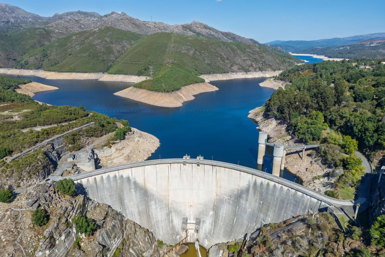 Vue aérienne d'un barrage voûte en béton retenant un lac aux berges dénudées, entre des montagnes rocheuses et boisées