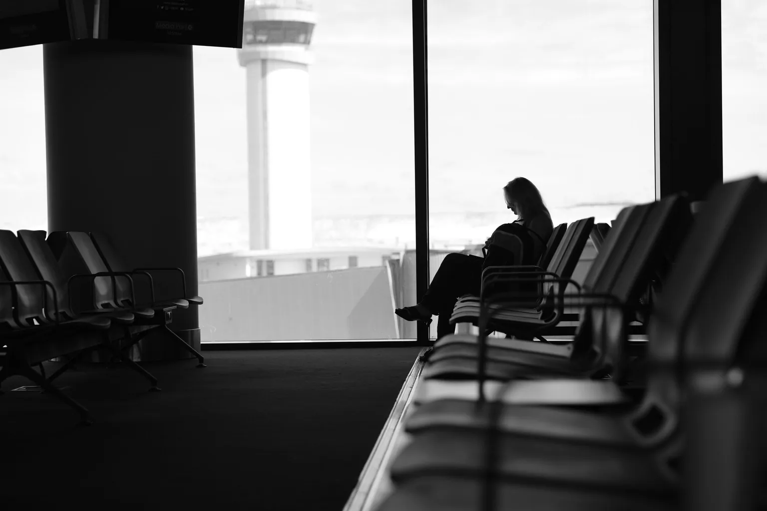 Silhouette d'une voyageuse assise dans une salle d'embarquement, devant une baie vitrée donnant sur une tour de contrôle