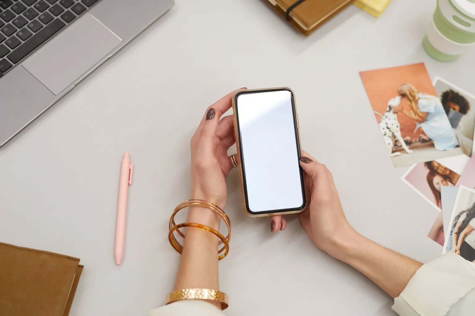 Mains tenant un smartphone à l'écran vierge sur un bureau clair, à côté d'un ordinateur portable et de photos imprimées