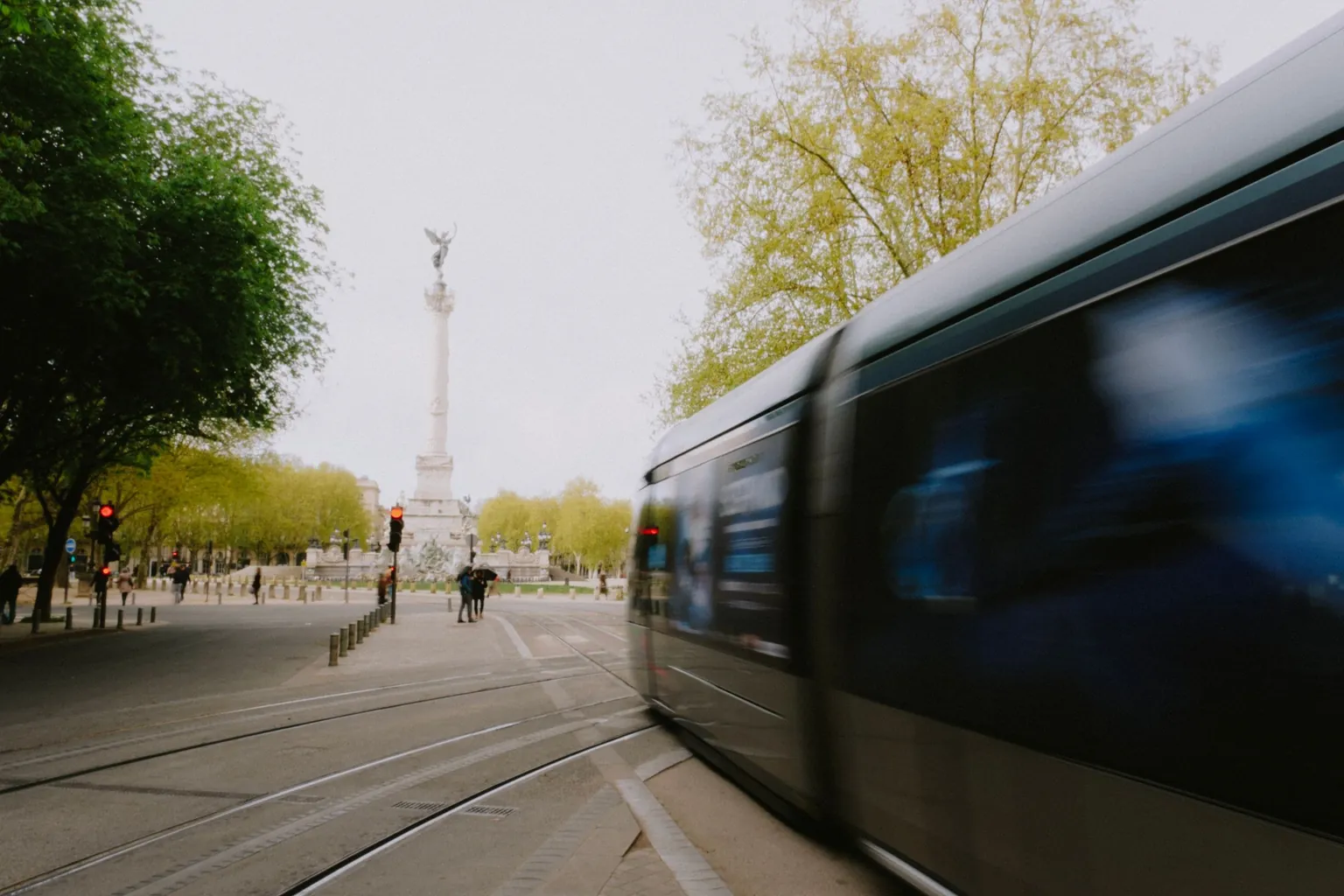 Tramway passant à vive allure devant une colonne monumentale surmontée d'une statue ailée, au bout d'une esplanade arborée