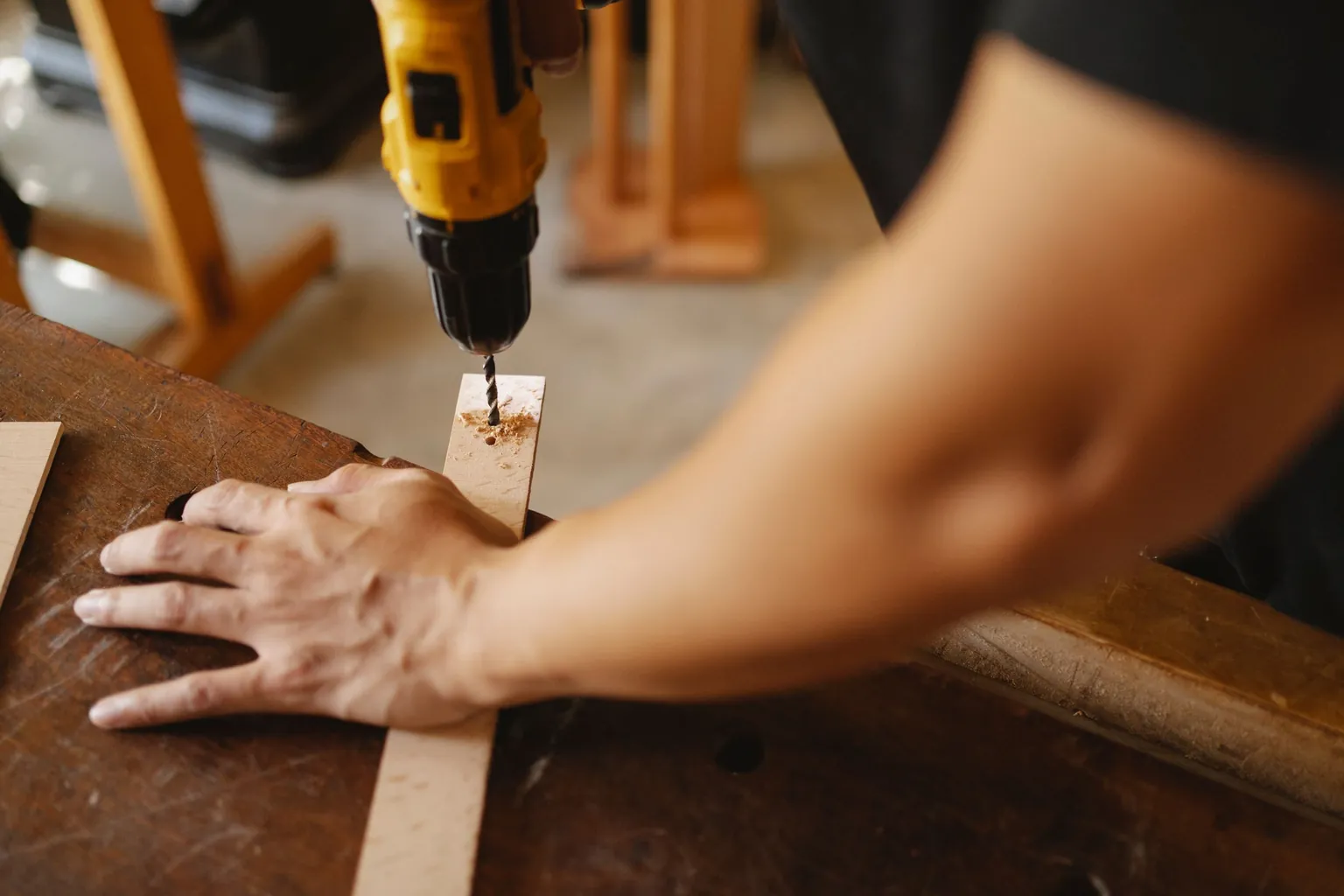Perceuse sans fil jaune perçant une latte de bois tenue à la main sur un établi