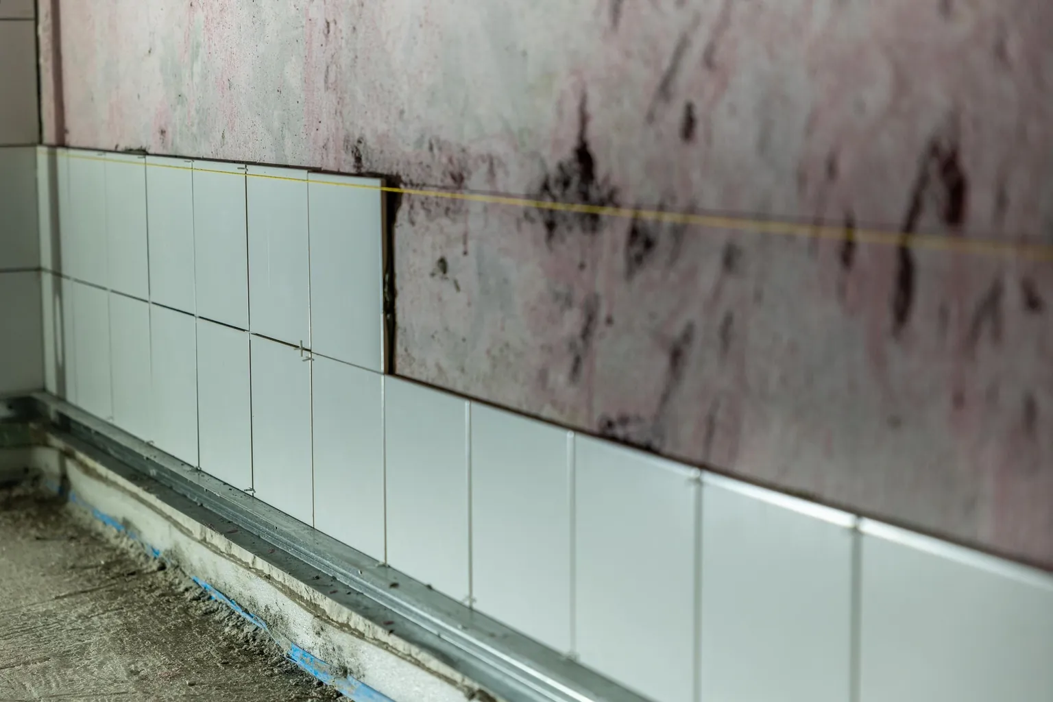 Carreaux de céramique blancs en cours de pose au bas d'un mur brut, avec cordeau de repère