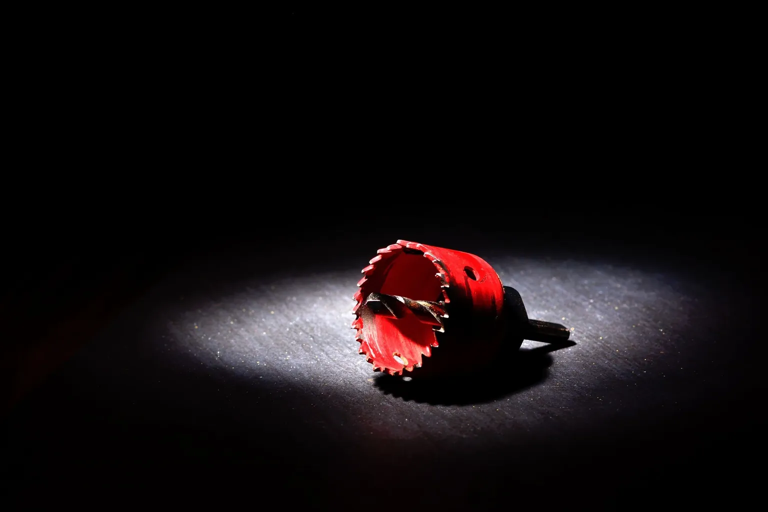 Scie cloche à denture rouge avec sa mèche de centrage, posée sous un faisceau de lumière sur fond sombre