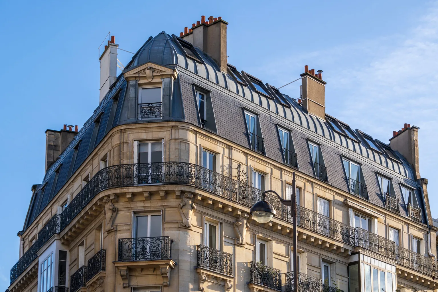 Angle d'un immeuble haussmannien en pierre claire, toit en zinc et balcons en fer forgé sous un ciel bleu