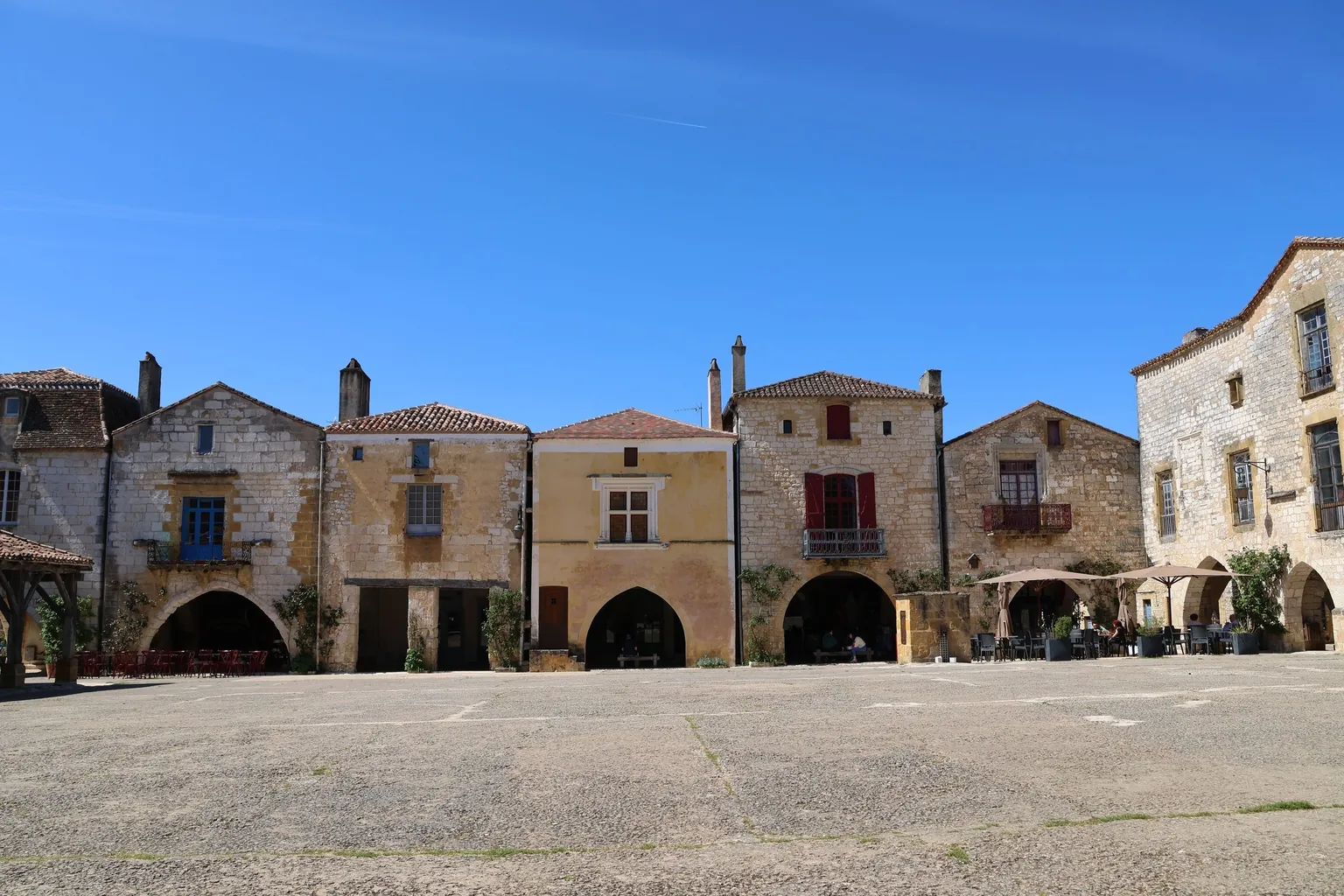 Place de bastide bordée de maisons de pierre à arcades sous un ciel bleu, terrasses sous les couverts